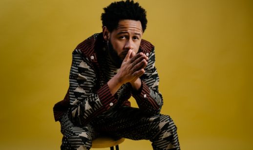 Emicida sentado com as mãos juntas próximas aos nariz em fundo amarelo