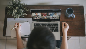 Pessoa de home office, com mesa com laptop em reunião remota, caderno, lápis e caneca de café