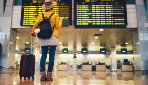 Pessoa de costas com mala de carrinho olhando o celular e o painel de voos, alusivo às viagens que beneficiam ações de Airbnb