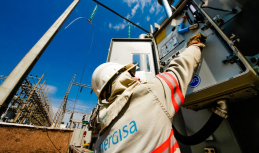 Funcionário da Energisa com uniforme, de costas, com destaque para o logo da empresa e torres de energia ao fundo, ângulo de baixo para cima