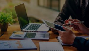 Close de mesa com laptop, papéis e bloco de anotações com duas pessoas conversando e olhando tablet, alusivo a um assessor de investimentos orientando cliente