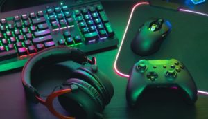 Teclado e mouse de computador, fone e controle de videogame, todos pretos e com detalhes iluminados, alusivo ao mercado de games