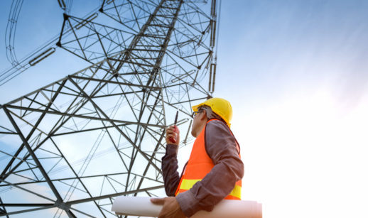 Perspectiva de baixo para cima de trabalhador com colete e capacete de proteção com rádio de comunicação nas mãos ao lado de torre de transmissão de energia, alusivo à privatização da Eletrobras