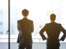 Dois executivos de terno preto, de costas, olhando por janela iluminada, alusivo ao Safra Report de outubro