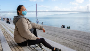 Mulher sentada com casaco bege, de máscara e óculos de sol, olhando a paisagem em Portugal