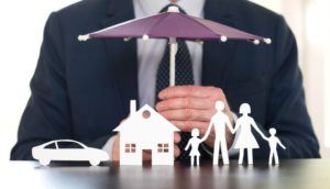 Pessoa segurando guarda-chuva de brinquedo pequeno sobre bonecas de famílias, casa e carro, alusivo ao seguro de vida