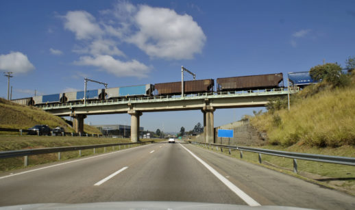 Vista de dentro de carro para trem sobre rodovia, alusivo ao avanço das ferrovias no Brasil