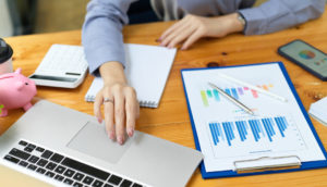 Mãos de duas pessoas sobre mesa bege com laptop e planilhas desenhadas em papel, alusivo à carteira administrada que tem gestão ativa