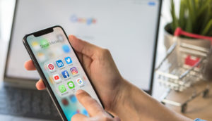 Pessoa segurando iPhone com a tela aberta na pasta de apps de redes sociais e laptop ao fundo desfocado aberto na página do Google, alusivo à proteção de dados pessoais