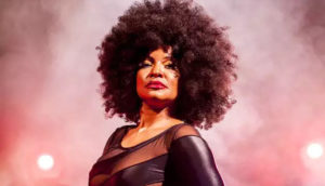 Elza Soares, de vestido preto, com fundo rosado