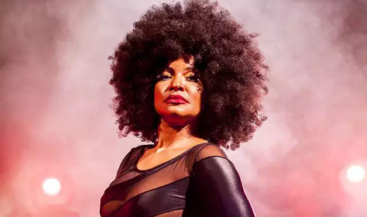 Elza Soares, de vestido preto, com fundo rosado