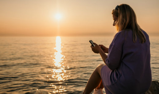 Mulher de costas sentada com celular em mãos e o mar com pôr do sol ao fundo, alusivo aos investimentos para janeiro