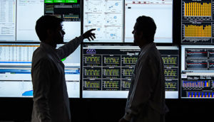 Silhueta de dois pesquisadores com um apontando para telas de computador atrás deles, alusivo às atividades da Alliar