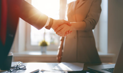 Homem e mulher em plano americano apertando as mãos com raio de sol entrando por janela ao fundo, alusivo às fusões e aquisições no Brasil em 2021