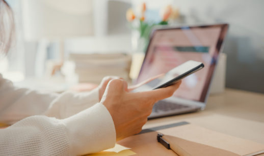 Mãos segurando e mexendo em celular com laptop ao fundo, alusivo ao vazamento de chaves do Pix