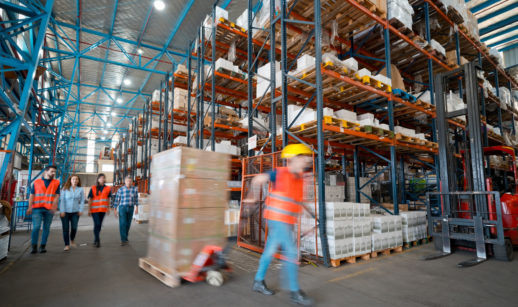 Armazém com estoques de produção, trabalhador com colete e capacete passando com carrinho de carregar produtos