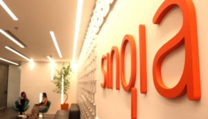 Interior de sala da Sinqia, com perspectiva de proximidade à parede dando destaque ao logo, à direita, em laranja