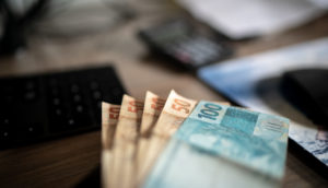 Notas de cem e cinquenta reais enfileiradas sobre mesa com calculadora e PC ao fundo, alusivo aos pedidos de transferência via sistema Valores a Receber, do Banco Central