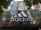 Fachada de loja da Adidas em prédio espelhado, com destaque para o logo em branco e árvores próximas nas laterais