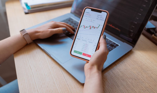 Close de mãos segurando celular com tela de gráficos de mercado e laptop aberto, alusivo ao investimento em commodities