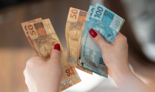 Mãos com unhas vermelhas pintadas contando notas de 50 e 100 reais, alusivo ao saque do FGTS