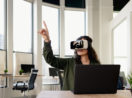 Mulher com óculos de realidade virtual na frente de laptop com mãe apontada para cima, alusivo à uma reunião no metaverso