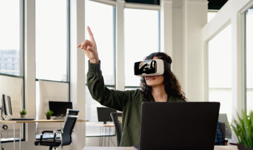 Mulher com óculos de realidade virtual na frente de laptop com mãe apontada para cima, alusivo à uma reunião no metaverso