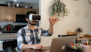 Homem de camisa xadrez azul com óculos de realidade virtual com laptop à frente, alusivo ao metaverso