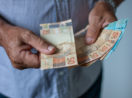 Close em mãos de uma pessoa com camisa azul contando notas de 50 e 100 reais, alusivo à restituição do IRPF 2022