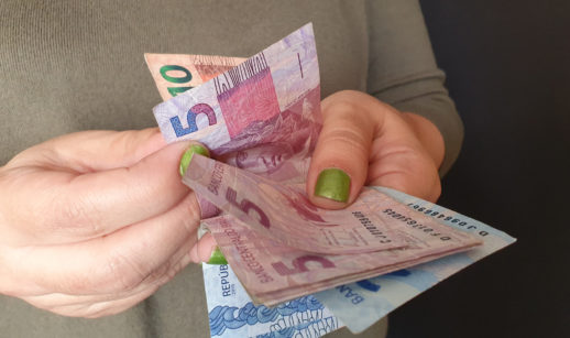 Mãos com unhas pintadas de verde contando notas de 5, 10 e 100 reais, alusivo aos valores a receber