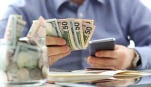 Homem tem um celular na mão e algumas notas de dinheiro, alusivo ao processo de comprar moeda estrangeira