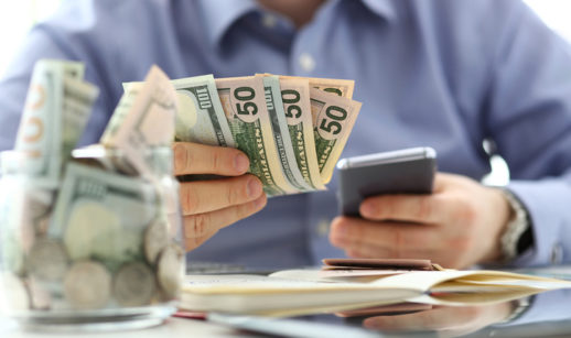 Homem tem um celular na mão e algumas notas de dinheiro, alusivo ao processo de comprar moeda estrangeira