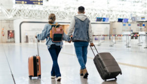 Um homem e uma mulher andam por um aeroporto com suas malas, em alusão a uma viagem de férias