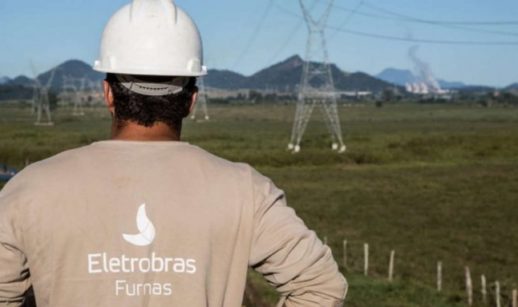 Funcionário de costas com roupa estampada 'Eletrobras Furnas' e capacete branco, em primeiro plano, e torres de transmissão de energia ao fundo