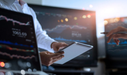 Close de telas de computador com gráficos do mercado financeiro e uma pessoa de camisa branca apontando para tablet, alusivo às top picks do Safra para a semana