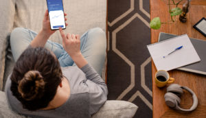 Perspectiva de cima para baixo de mulher sentada em sofá mexendo no celular, estudando o que é o investimento em COE, com café, fone de ouvido, caderno e caneta sobre mesa à sua direita