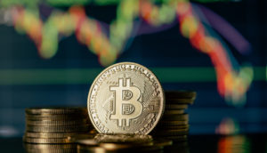 Representação do Bitcoin em forma física, com destaque para o logo da criptomoeda e gráficos de sistema financeiro atrás