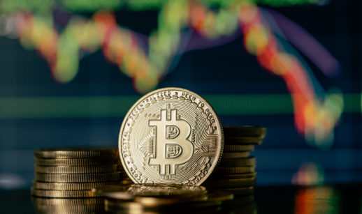 Representação do Bitcoin em forma física, com destaque para o logo da criptomoeda e gráficos de sistema financeiro atrás