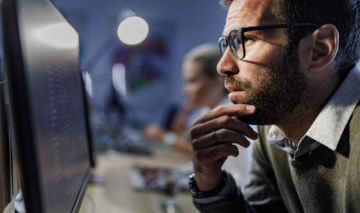 Homem de barba e óculos, de perfil, com mão no queixo, olhando para o computador, alusivo ao escritório de investimentos