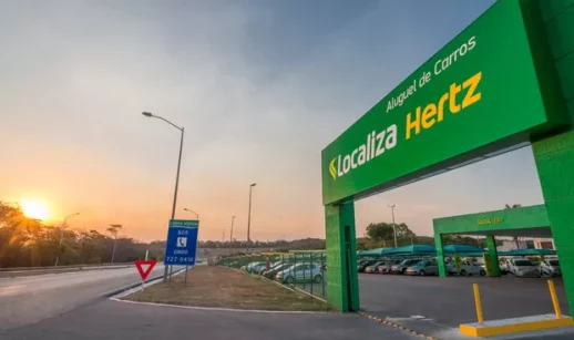 Entrada de pátio da Localiza (RENT3), com frota de carros parada dentro ao lado de rodovia com pôr do sol