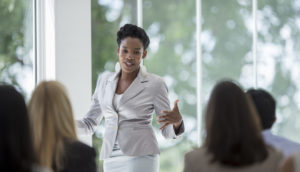 Mulher na liderança de reunião de blazer rosa claro com saia branca gesticulando de frente para mulheres sentadas