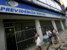 Fachada de agência da Previdência Social, que concede benefícios do INSS, com três pessoas passando na frente na calçada