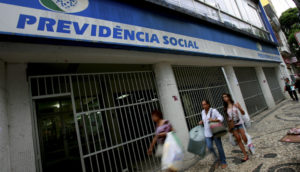 Fachada de agência da Previdência Social, que concede benefícios do INSS, com três pessoas passando na frente na calçada