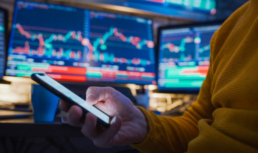 Close de pessoa com suéter amarelo mexendo em celular com telas de PC atrás, alusivo às operações em renda variável