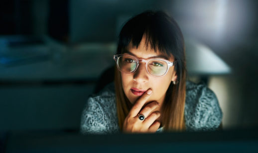 Mulher de óculos de armação de plástico transparente e franja olhando para tela de computador, alusivo ao pensamento nos investimentos por perfil de investidor