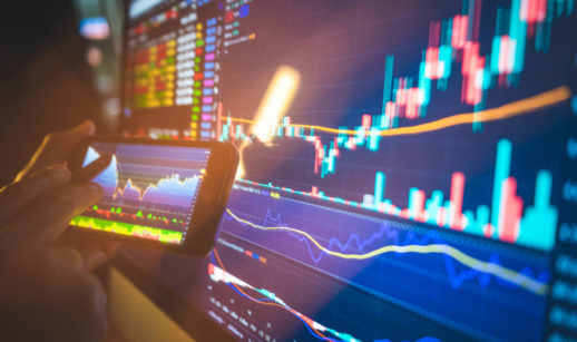 Close de telas com gráficos do mercado financeiro em celular e computador, alusivo à oscilação das ações do Hapvida