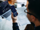 Pessoa analisando gráficos financeiros em um smartphone, com laptop exibindo gráficos e documentos ao fundo