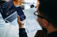 Pessoa analisando gráficos financeiros em um smartphone, com laptop exibindo gráficos e documentos ao fundo