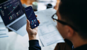 Pessoa analisando gráficos financeiros em um smartphone, com laptop exibindo gráficos e documentos ao fundo