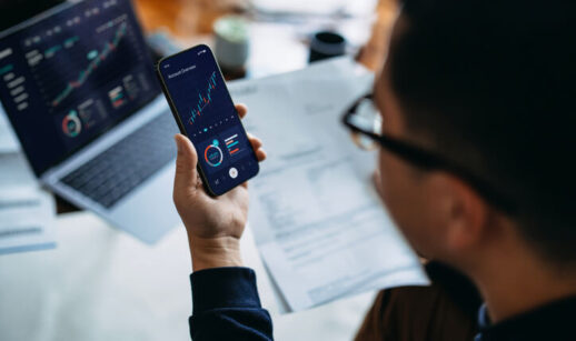 Pessoa analisando gráficos financeiros em um smartphone, com laptop exibindo gráficos e documentos ao fundo